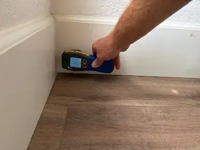 Hand holding a moisture meter against the bottom of a wall above a wood floor